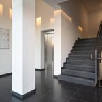 Haupteingang mit Treppe und Aufzug. Weiße Wände und dunkler Boden.