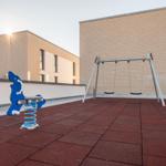 Kinderspielplatz mit Schaukel und blauem Feder-Spielgerät auf rotem Boden.