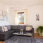 Ein Wohnzimmer mit grauem Sofa, Glas-Tisch und Pflanze. Fenster mit Vorhängen.