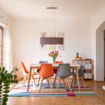 Esszimmer mit Holztisch, bunten Stühlen und buntem Teppich. Fenster, Pflanze und Wandbilder sichtbar.