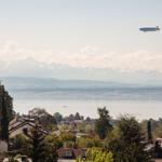 Luftschiff fliegt über See und Berge mit Blick auf Wohnhäuser.