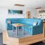 Ein Wohnbereich mit blauem Sofa, Holzpodest und Küchenbereich im Hintergrund.