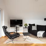 Ein Wohnzimmer mit schwarzem Sofa, Sessel und Fernseher. Holzboden und große Fenster.