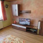 Wohnzimmer mit Fernseher, Schrank und Holzboden. Fenster mit Vorhängen.