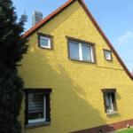 Gelbes Haus mit braunen Fensterrahmen und rotem Dach unter blauem Himmel.