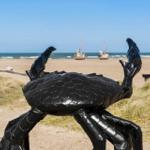 Eine schwarze Krabbenstatue am Strand mit Booten im Hintergrund.