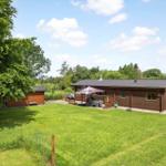 Holzhaus mit Garten, Terrasse und Sonnenschutz. Links Nebengebäude, rechts Parkplatz.