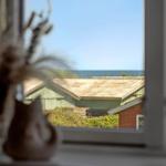 Durch das Fenster ist das Meer und ein Haus mit Wellblechdach zu sehen.