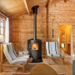 Holzhaus mit Kamin, zwei Sesseln und Fenster. Tür führt in angrenzenden Raum.