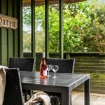 Terrasse mit Tisch, Stühlen und einer Flasche Wein. Hintergrund: grüne Vegetation.