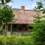 Haus mit strohgedecktem Dach und roter Tür, umgeben von Bäumen und Grünflächen.