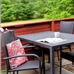 Terrasse mit Tisch, Stühlen und Kissen. Hintergrund: grüne Bäume und rote Holzablage.