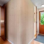 Haus mit Holzwänden, Eingangstür und Blick auf Grün. Im Hintergrund ist ein schwarzer Sessel sichtbar.