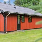 Rotes Holzhaus mit schwarzem Dach und Fenstern. Grasfläche und Bäume im Hintergrund.
