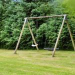 Hinter einem grünen Heckenzaun steht ein Holzschaukelstand mit zwei Schaukeln auf einer Wiese.