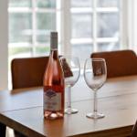 Eine Flasche Roséwein und zwei Gläser stehen auf einem Holztisch. Im Hintergrund sind Stühle und Fenster zu sehen.