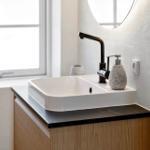 Waschbecken mit Wasserhahn und Seifenspender auf Holzschrank. Hintergrund: Fenster und runder Spiegel.