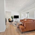 Wohnzimmer mit braunem Sofa, Fernseher und Holzboden. Blick in die Küche.