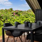 Terrasse mit Tisch, Stühlen und Teekanne vor blühenden Sträuchern.