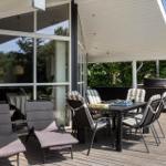 Terrasse mit Tisch, Stühlen und Liegestühlen. Hintergrund: Glasfassade und grüne Bäume.