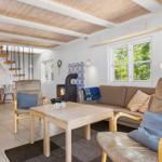Wohnzimmer mit Sofa, zwei Sesseln, Tisch und Kaminofen. Holzdecke und Fenster mit Blick auf Grün.