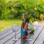 Auf einem Tisch stehen zwei Weingläser und eine Flasche alkoholfreies Rosé. Im Hintergrund Blumen und Grün.