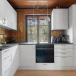 Küche mit weißen Schränken, dunkler Arbeitsplatte und Holzwand. Fenster mit Blick auf Wald.
