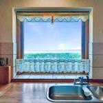 Kitchen with view of green field and sky. Stainless steel sink, wooden countertop, and window with curtain.