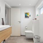 Badezimmer mit Waschtisch, Toilette und Fenster. Wandbild und runder Spiegel sichtbar.
