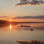Sonnenuntergang über ruhigem See mit Reflexion und Felsen im Vordergrund.