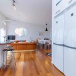 Küche mit weißen Schränken und Kühlschrank. Wohnbereich mit braunem Sofa und runden Fenster.