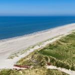 Weiterer Blick auf den Strand mit Dünen und dem Meer unter blauem Himmel.