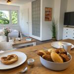 Esstisch mit Brot und Kaffee, Wohnbereich mit Fernseher und Fenster.