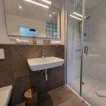 Badezimmer mit Dusche, Waschbecken und Spiegel. Toilette und Handtuch sind sichtbar.