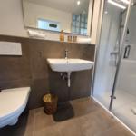 Badezimmer mit Dusche, Waschbecken und Toilette. Wand und Boden sind mit braunen Fliesen ausgelegt.