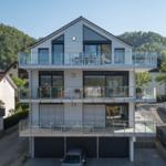 Moderne Villa mit drei Terrassen und großem Balkon. Garagenbereich und grüne Hügel im Hintergrund.