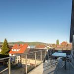 Terrasse mit Tisch und Stühlen, Blick auf Dächer und See unter blauem Himmel.