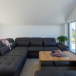 Ein Wohnzimmer mit einem schwarzen Sofa, einem Holztisch und großen Fenstern.