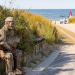 Holzfigur eines Mannes am Strandweg mit Dünen und Meer im Hintergrund.