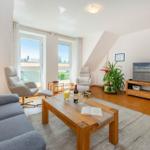 Wohnzimmer mit Sofa, zwei Sesseln, Holztisch und Fernseher. Fenster mit Blick nach draußen.