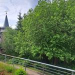 Weg mit Metallgeländer führt durch grüne Bäume zu einem Turm mit Zwiebelhaube.