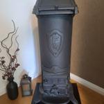 Schwarzer Kachelofen mit Wappen auf Holzboden neben Vase und Laterne.