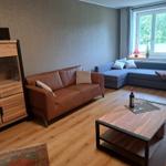 Wohnzimmer mit braunem Ledersofa, blauem Ecksofa, Holztisch und Fenster mit Blick auf Bäume.