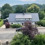 Haus mit grauem Dach und Garten, umgeben von Bäumen und einem Straßenbereich.