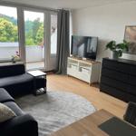 Wohnzimmer mit Sofa, Fernseher und Balkonblick auf grüne Bäume.