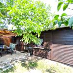 Terrasse mit Sitzgruppe, Grill und Schatten unter Bäumen vor einem Holzhaus.