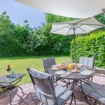 Terrasse mit Tisch, Stühlen und Sonnenschirm. Hintergrund: grüner Garten und Hecke.