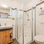 Badezimmer mit Dusche, Waschbecken und Toilette. Holzunterschrank und Spiegel mit Beleuchtung.