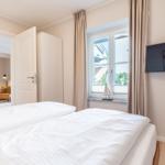 Schlafzimmer mit Bett, Fenster und TV. Durchgang zur Wohnfläche mit Sofa.