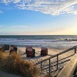 Strand mit Liegestühlen und Holzsteg. Sonne geht unter. Wellen brechen am Horizont.